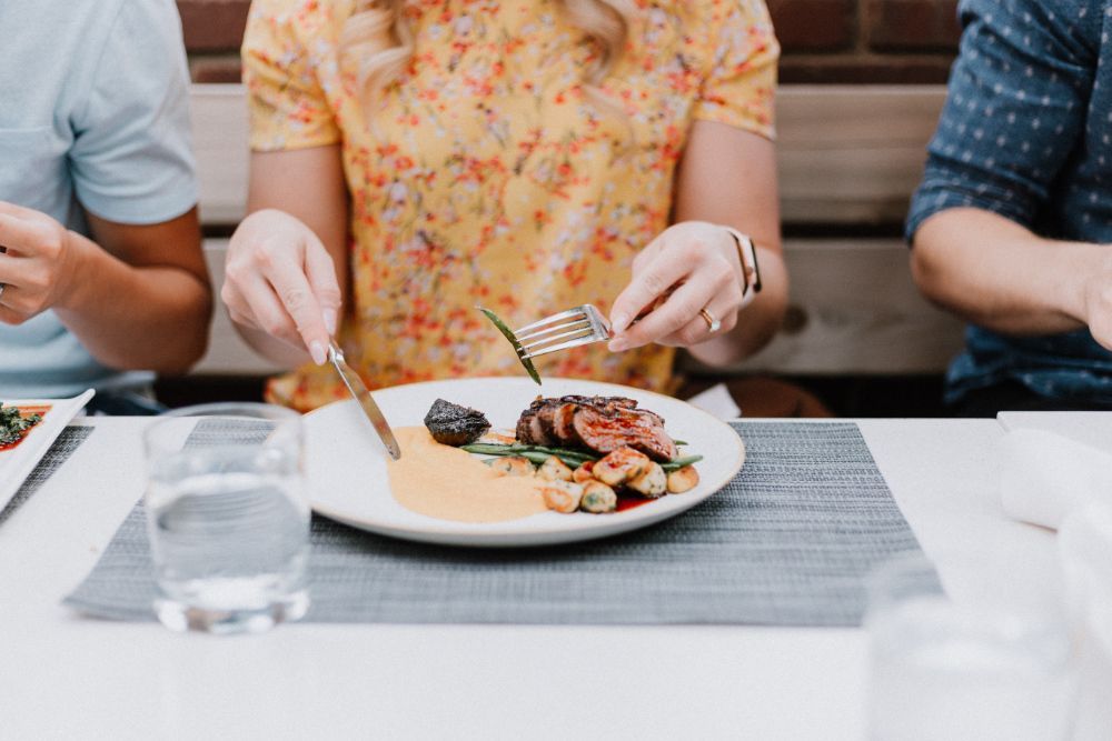 Lady in yellow dress with floral pattern eating steak and vegetables in a restaurant.